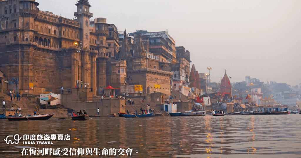 這張圖片拍攝自印度瓦拉納西（Varanasi）的恆河（Ganges River）岸邊，畫面中可見古老的石建寺廟、樓房、寬大的台階與停泊的木船，呈現出宗教與日常生活交織的獨特場景。清晨的薄霧與暖色燈光交融，讓整體氛圍充滿神聖與寧靜，呼應下方文字「在恆河畔感受信仰與生命的交會」，傳達出印度宗教文化的深度與情感張力。