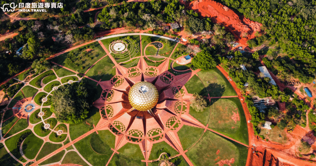 這是一張俯瞰印度南部曙光之城（Auroville）的航拍照片，畫面中央為象徵人類和諧與靈性成長的「母親殿堂」（Matrimandir），金色圓球在陽光下閃耀，周圍環繞對稱的花瓣狀紅磚步道與綠地。這裡被稱為「靈性烏托邦」，吸引世界各地遊客與修行者前來體驗沉思與社群生活。左下角可見「印度旅遊專賣店 India Travel」標誌，強化品牌識別。