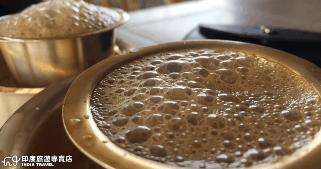 這張圖片展示了南印度經典的濾泡咖啡（Filter Coffee），盛裝在傳統的金屬杯與碟中。泡沫綿密的咖啡表面，散發出濃厚香氣，帶有獨特的奶香與烘焙風味。這種咖啡是南印度日常生活的重要部分，常搭配早餐或午後小點，象徵著當地的飲食文化與悠閒氛圍。