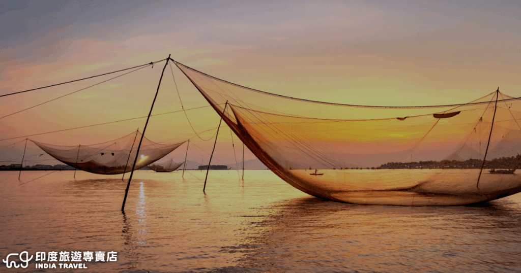 夕陽下的柯欽（Fort Kochi）海岸，中國漁網（Chinese Fishing Nets）隨風搖曳，漁夫的小舟點綴在水面上，天空被染成橙紅與紫色的漸層。這個場景是南印度喀拉拉邦（Kerala）的代表性畫面，結合漁業文化與浪漫的海岸氛圍，常被視為到訪柯欽必拍的經典景點之一。左下角標示「印度旅遊專賣店 India Travel」字樣，呈現專業品牌識別。