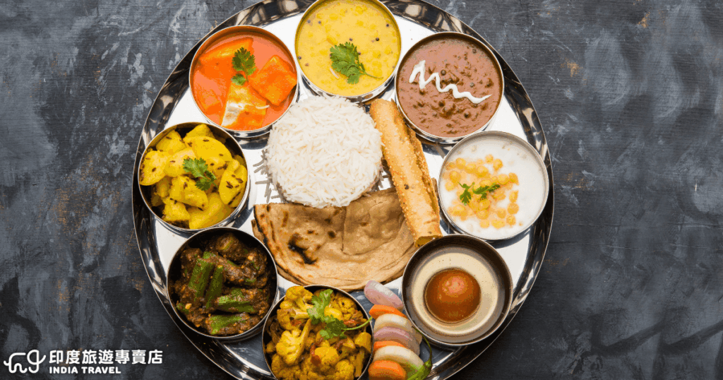 這張圖片展示印度經典的傳統餐盤 Thali,中央擺放著一碗白米飯,周圍環繞著多種小碗料理,包括咖哩、扁豆湯(Dal)、炒蔬菜、優格、甜點與餅類(如Chapati或Roti)。每一道菜色色彩鮮豔,口味兼具酸、甜、辣與香料風味,象徵著印度飲食文化中「多元共融」的精神。Thali 不僅是日常餐食的一部分,也是體驗印度飲食文化的重要方式,讓人一盤就能嚐遍印度的豐富滋味。
