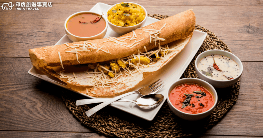 這張圖片展示的是南印度經典料理「Masala Dosa」，一種由米漿與扁豆發酵製成的酥脆薄餅，內餡通常包裹著香料調味的馬鈴薯泥。旁邊搭配多種醬料（chutney），包含椰子醬、番茄醬和扁豆湯（Sambar），風味層次豐富。Masala Dosa 是南印度日常早餐與小吃的代表，酥脆外皮與滑順餡料的結合，讓它成為旅人必嚐的在地美食之一。