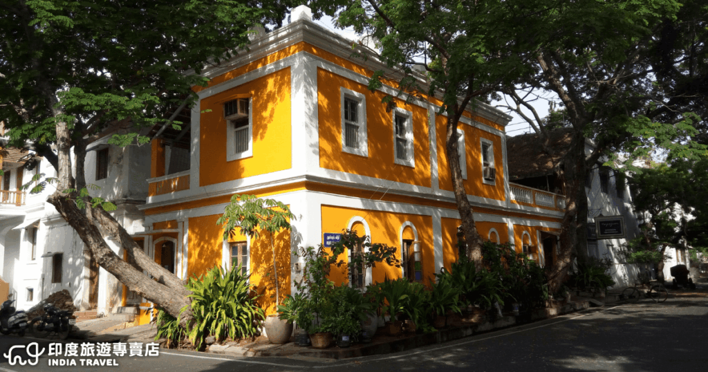 圖片展現南印度朋迪榭里（Pondicherry）的法式街區景象，一棟鮮豔亮眼的黃色殖民風格建築坐落在綠蔭環繞的街角，白色線條勾勒出典雅的外觀。周圍的植栽與樹木在陽光下投下斑駁的光影，營造出悠閒浪漫的氛圍。朋迪榭里因為過去的法國殖民歷史，被稱為「印度的法國小鎮」，融合了歐洲建築與印度文化特色，是南印度旅遊的重要亮點。