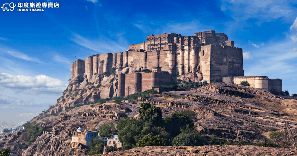 照片展示印度拉賈斯坦邦焦特布爾（Jodhpur）的梅蘭加爾岩堡（Mehrangarh Fort），坐落在高達約122公尺的山丘上，整座堡壘以紅砂岩建造，牆壁厚實而壯觀。堡壘內部有宮殿、庭院與博物館，保存了精美的壁畫與鏤空石雕。這裡曾是馬爾瓦爾王朝的重要權力中心，如今已成為印度最壯觀的古堡之一，也是《蝙蝠俠：黑暗騎士》、《泰山傳奇》等電影的取景地。藍天與岩石地形襯托出古堡的雄偉氣勢，左下角有「印度旅遊專賣店 India Travel」標誌，適合作為印度拉賈斯坦歷史文化旅遊的形象圖。