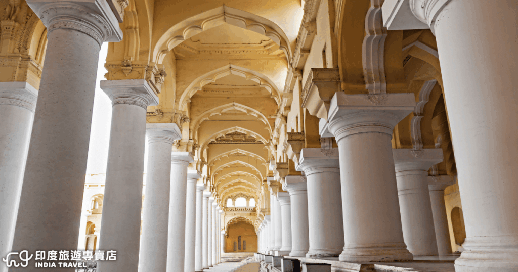 畫面展示印度泰米爾納德邦（Tamil Nadu）馬杜賴（Madurai）的納亞克宮殿（Thirumalai Nayak Palace）拱廊內部。高聳的白色圓柱與層層堆疊的黃金色拱門，呈現壯麗的德拉威風格與伊斯蘭建築元素融合。此宮殿建於17世紀，是納亞克王朝最具代表性的宮殿之一，曾經作為皇宮和政務中心。光影灑落在迴廊間，讓建築顯得更加莊嚴與宏偉。左下角有「印度旅遊專賣店 India Travel」標誌，適合用於文化建築與歷史旅遊的宣傳內容。