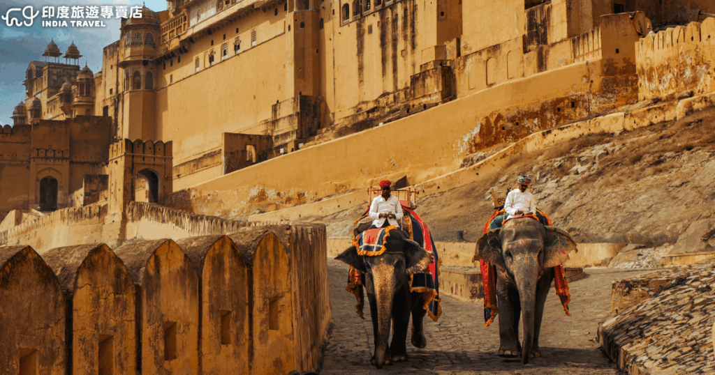 畫面中展現拉賈斯坦邦斋浦尔（Jaipur）的琥珀堡（Amber Fort）入口，兩位身穿傳統服飾的馴象師正騎著裝飾華麗的大象緩緩走向城門，象徵當地旅遊的文化體驗。金黃色城牆映襯下，氛圍既莊嚴又具異國情調，是遊客到訪琥珀堡的經典畫面之一。