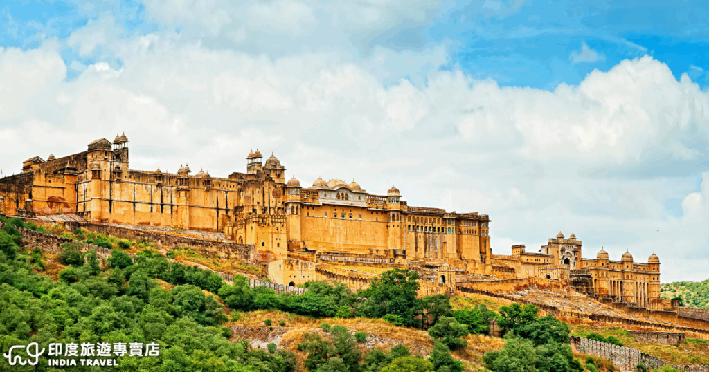 這張圖片呈現印度齋浦爾（Jaipur）的琥珀堡（Amber Fort）全景，金黃色砂岩建築依山而建，城牆延伸至山坡，與藍天白雲和綠意相互映襯，展現拉賈斯坦邦皇室的歷史風華與建築之美。