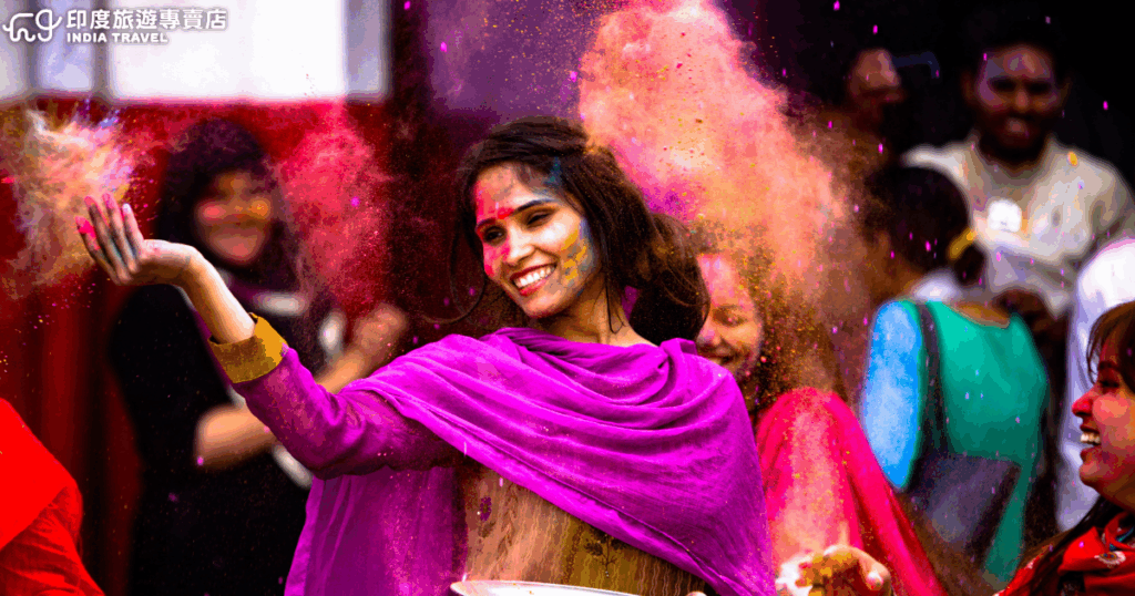 畫面捕捉印度灑紅節（Holi Festival）中女子揮灑彩粉的瞬間，她身穿亮紫色傳統披肩，臉上與空氣中瀰漫著橙、粉、紫等彩粉，露出開朗笑容，象徵節慶中喜悅與祝福的氛圍。背景可見群眾同樣歡笑互動，整體構圖色彩飽和、動感十足，展現印度節慶的熱情與人文魅力，非常具感染力與文化象徵性。