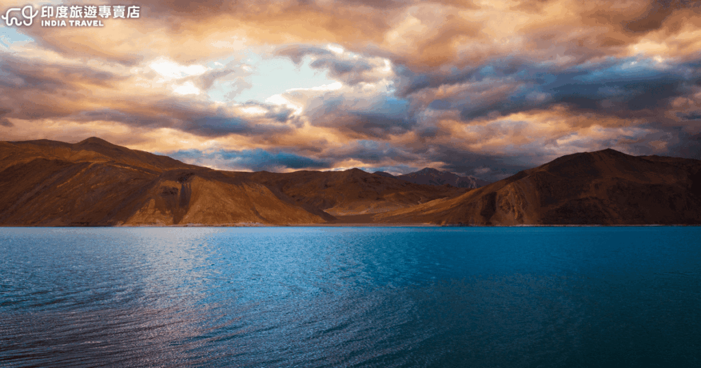 這張圖片呈現位於印度拉達克（Ladakh）的班公錯湖（Pangong Lake）黃昏景色。湖面呈現深藍色，映照著遠方荒蕪山脈的輪廓，天空覆蓋著層層厚實雲層，夕陽的橘金色光線穿透雲隙，讓整個景象帶有靜謐又壯觀的氛圍。此圖片非常適合用於印度北部旅遊介紹、拉達克旅遊資訊、自然景觀推薦或高原湖泊的文章主視覺。